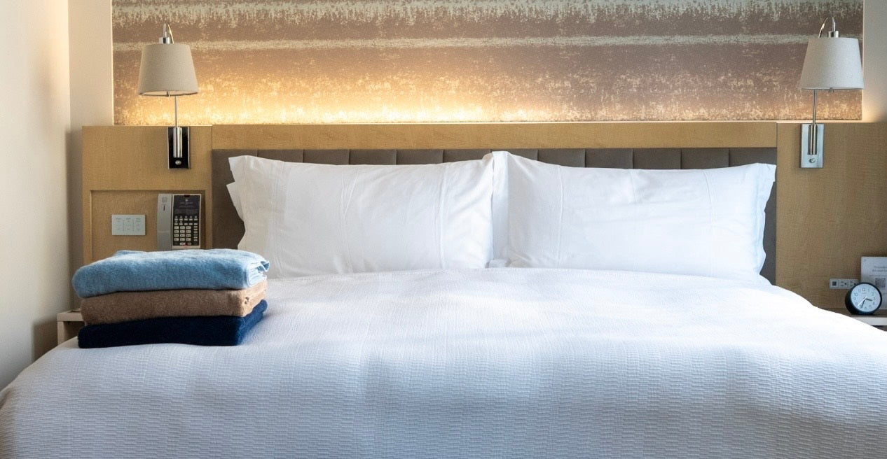 Neatly made bed with white bedding and folded towels in a well-lit bedroom.