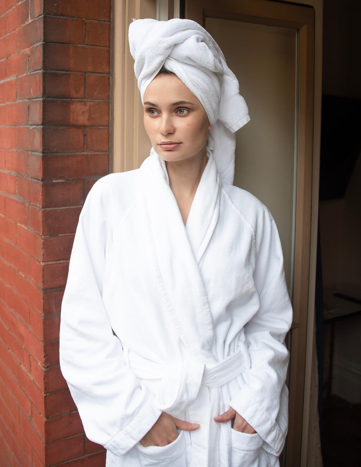 Woman wearing a white robe and towel wrapped around her head, standing against a brick wall.